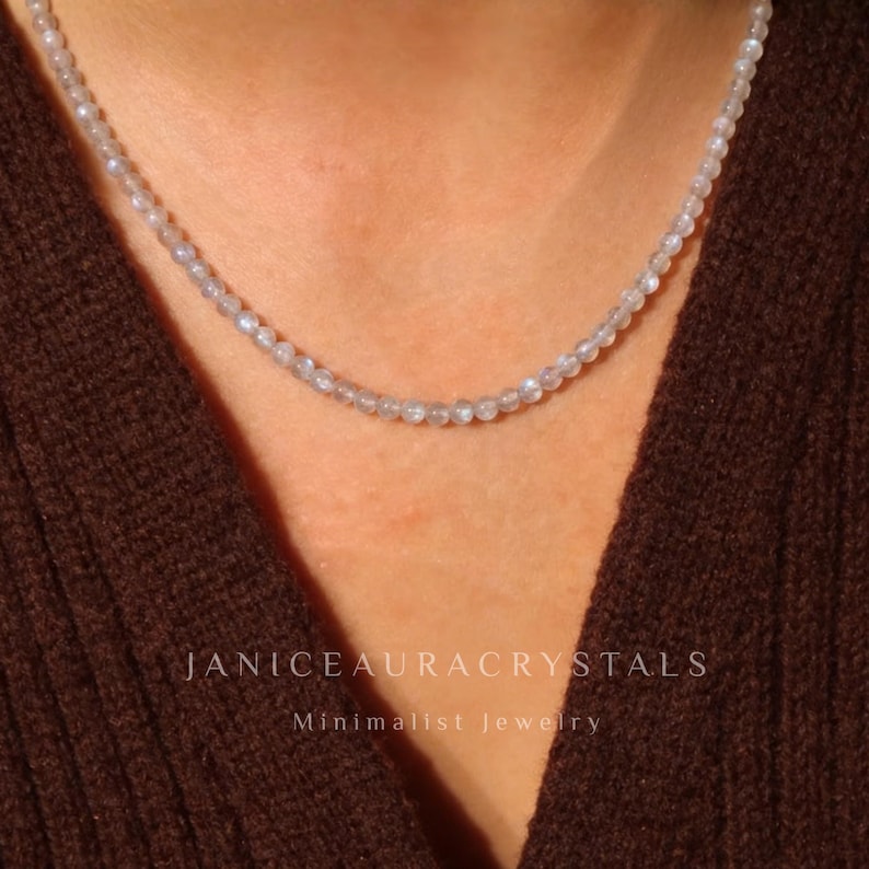 May include: A delicate necklace composed of small, round, light-colored beads. The necklace is displayed against a brown background. The text "JANICEAURACRYSTALS" and "Minimalist Jewelry" are visible.