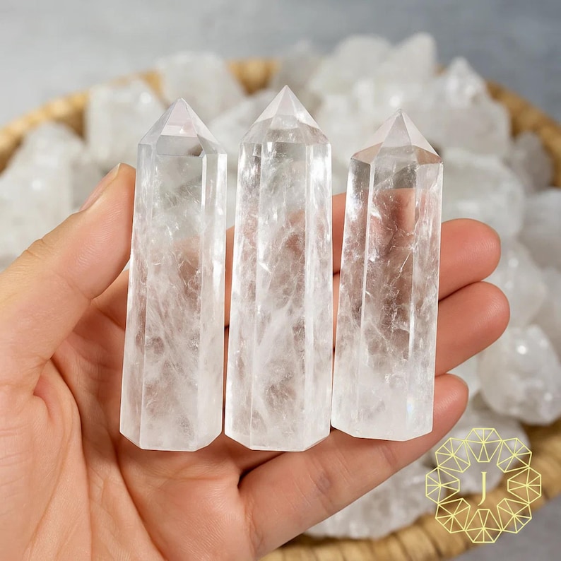 May include: Three clear quartz crystal points with a hexagonal base are held in a hand. The crystals are transparent with internal veining. A basket of raw quartz crystals is in the background. The crystals are a light, neutral colour.