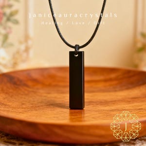 May include: A black rectangular pendant necklace suspended from a black cord. The pendant is smooth and polished. The image includes the text "janiceauracrystals" and "Healing / Love / Gift". The necklace rests on a wooden plate.
