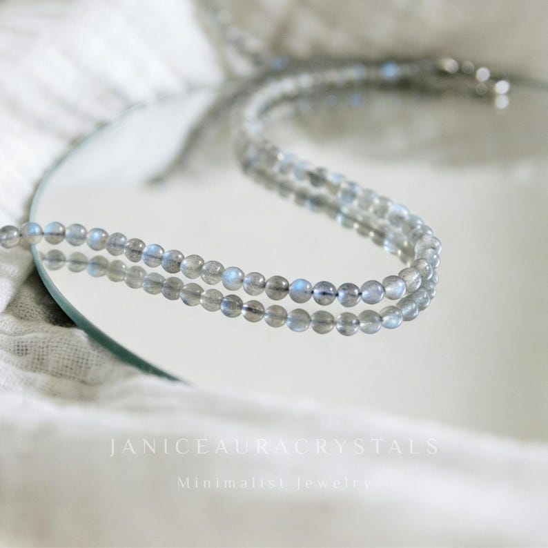 May include: A minimalist necklace featuring small, round, gray beads with blue iridescence. The necklace is displayed on a reflective surface, with the text "JANICEAURACRYSTALS" and "Minimalist jewelry" in white.