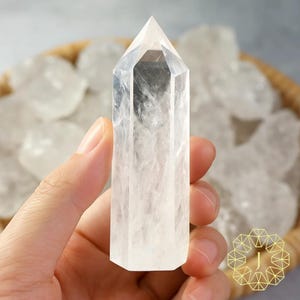 May include: A clear quartz crystal point held in a hand. The crystal is transparent with internal veining and a pointed top. Other quartz crystals are in a woven basket in the background. The crystal is a natural, polished gemstone.