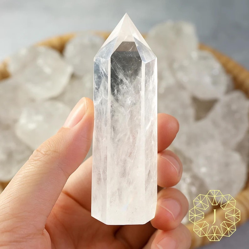May include: A clear quartz crystal point held in a hand. The crystal is transparent with internal veining and a pointed top. Other quartz crystals are in a woven basket in the background. The crystal is a natural, polished gemstone.