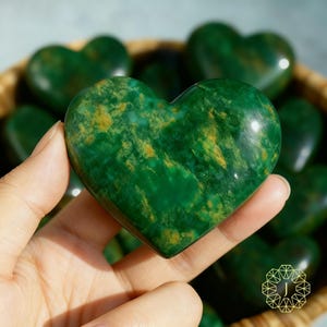 May include: A polished green heart-shaped stone with yellow and gold veining. The stone is held in a hand, with other heart-shaped stones visible in the background. The stones are in a basket. The heart-shaped stones are a decorative item.