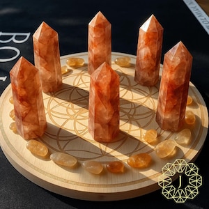 May include: Six orange-red crystal towers and several smaller tumbled stones are arranged on a wooden, circular base with a geometric design. The crystals have a translucent appearance, and the base is light brown.
