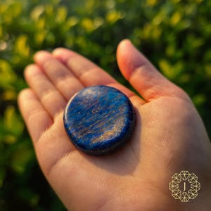 May include: A round, blue stone with gold flecks is held in an open hand. The stone has a polished surface and a dark blue color with streaks of lighter blue and gold. The background is blurred green foliage.
