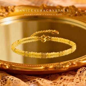 Op de afbeelding: Een delicate gele kralenarmband met gouden accenten, liggend op een reflecterend oppervlak. De armband heeft kleine, ronde gele kralen en goudkleurige metalen kralen. De tekst "JANICEAURACRYSTALS handicrafts / Love / Gift" is zichtbaar.