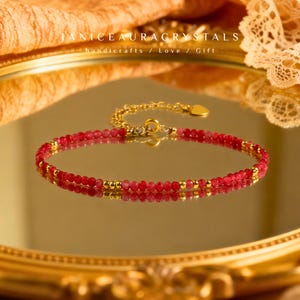 May include: A delicate red beaded bracelet with gold accents. The bracelet is displayed on a reflective surface, with the text "JANICEAURACRYSTALS handicrafts / Love / Gift" visible in the background. The bracelet's design features small, round red beads and gold-colored beads.