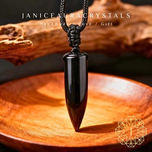 Puede incluir: Un collar con colgante de cristal de obsidiana negra. El colgante tiene forma alargada y pulida, con un extremo puntiagudo, suspendido de un cordón trenzado negro. El collar descansa sobre un plato de madera. El texto "JANICEAURACRYSTALS" es visible.