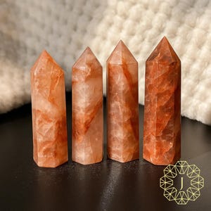 May include: Four orange-red quartz crystal towers with pointed tops. The crystals have a translucent appearance with reddish-orange veining. They are arranged in a row on a dark surface, with a soft, white background.