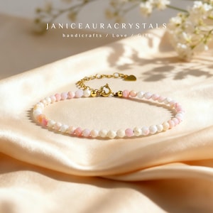 May include: A delicate bracelet featuring small, round, pink and white beads strung together. The bracelet has a gold-colored clasp and chain, with the text "JANICEAURACRYSTALS handicrafts / Love / Gift" in the background.