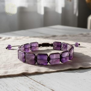 May include: A bracelet featuring rectangular amethyst stones strung on a dark cord. The purple stones are set in a square pattern, with a small amethyst bead at each end of the cord. The bracelet rests on a neutral-colored fabric.