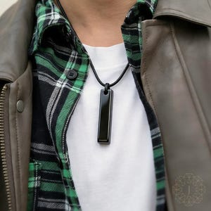 May include: A black rectangular pendant necklace. The pendant is suspended from a black cord. The person is wearing a white t-shirt, a green and black plaid shirt, and a gray leather jacket.