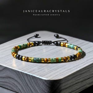 May include: A beaded bracelet with green, brown, black, and gold-colored beads. The bracelet is adjustable with a black cord and features the text "JANICEAURACRYSTALS Handcrafted Jewelry".