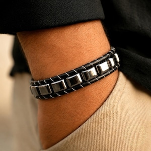 May include: A black leather bracelet featuring a row of polished, rectangular metallic beads. The beads are secured between two parallel black leather cords, which are joined by thin, light-colored stitching that wraps around the leather.