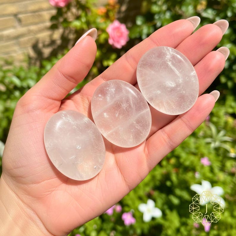 May include: Three oval-shaped rose quartz stones are displayed in a hand. The stones are a soft pink color and have a translucent quality. The background features green foliage and pink flowers, suggesting a natural setting.