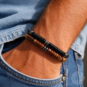 May include: Two beaded bracelets. One bracelet is black, the other is brown. Both bracelets have silver-colored metal accents. The bracelets are worn on a wrist, with a blue denim background.