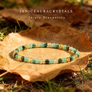 May include: A beaded bracelet with green, yellow, and clear gemstones. The bracelet is resting on a brown leaf. The text "JANICEAURACRYSTALS" and "Triple Prosperity" are at the top of the image.