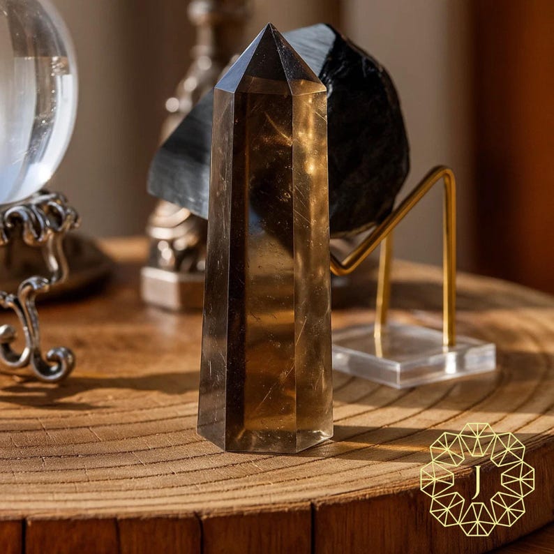 May include: A tall, faceted smoky quartz crystal point. The crystal is a translucent brown color and stands upright on a wooden surface. Other crystals and decorative objects are in the background.
