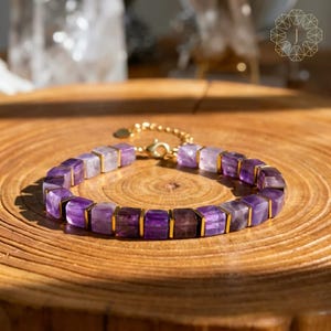 Puede incluir: Un brazalete con cuentas cuadradas de amatista y detalles metálicos dorados. El brazalete se exhibe sobre una superficie de madera, resaltando la combinación de colores púrpura y dorado. El cierre es dorado.