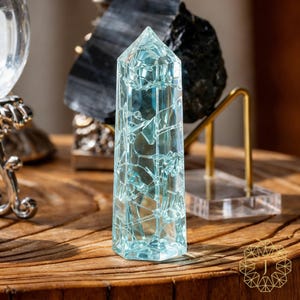 May include: A tall, light blue crystal obelisk with a fractured, geometric design. The crystal is set on a wooden surface, with other decorative objects in the background. The obelisk is approximately 6 inches tall.
