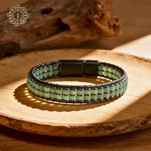 May include: A black leather bracelet with rows of light green beads and silver accents. The bracelet has a magnetic clasp and is displayed on a wooden plate. The bracelet is a piece of jewelry.