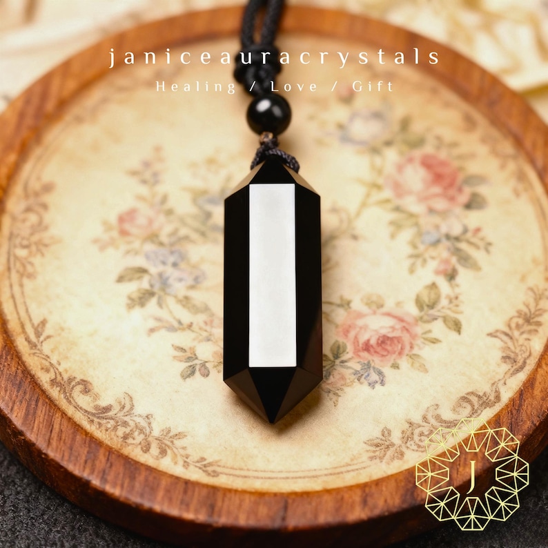 May include: A black and white crystal pendant necklace. The pendant is a hexagonal shape with a vertical white stripe. The necklace is on a floral patterned tray with the text "janiceauracrystals Healing / Love / Gift".