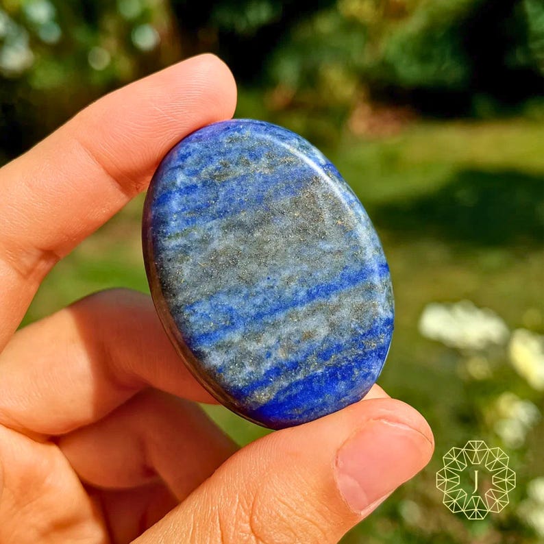 May include: A polished, oval-shaped lapis lazuli stone. The stone features a deep blue color with streaks of gray and gold. The gemstone is held in a hand, with a blurred green background.