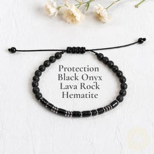 May include: A beaded bracelet featuring black lava rock beads, cylindrical black onyx, and metallic hematite spacers. The adjustable cord has a sliding knot closure with two small black beads at the ends. Text reads: Protection Black Onyx Lava Rock Hematite.