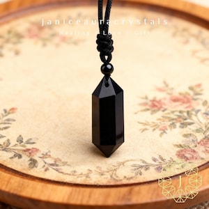 May include: A black obsidian crystal pendant necklace. The pendant is a faceted, elongated hexagonal shape, suspended from a black cord. The background features floral patterns and the text "janiceauracrystals Healing Love Gift".