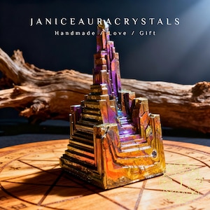 May include: A colorful bismuth crystal sculpture with a layered, geometric design. The crystal features iridescent hues of gold, purple, and blue. The text "JANICEAURACRYSTALS Handmade / Love / Gift" is displayed above the crystal.