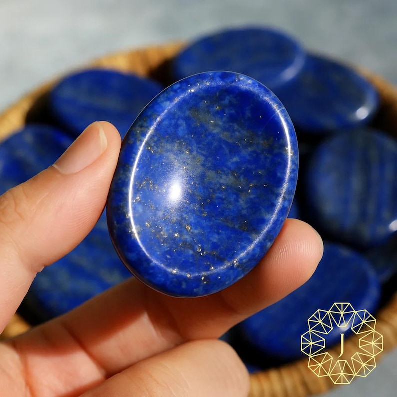 May include: A hand holding a polished, oval-shaped lapis lazuli stone. The stone is a deep blue with gold flecks and a smooth, reflective surface. Other similar stones are in a woven basket.