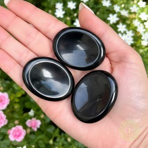 May include: Three polished, oval-shaped black obsidian stones. The stones are smooth and reflective, with a glossy finish. The stones are held in a hand, with a blurred background of flowers and greenery.