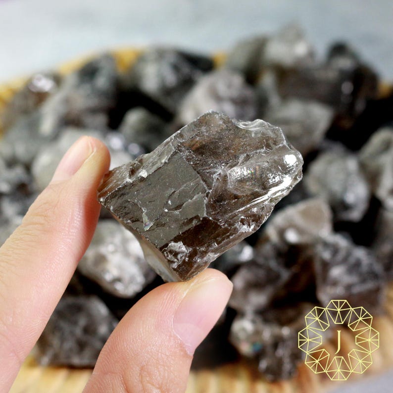 May include: A hand holding a rough, translucent, smoky quartz crystal. The crystal is a dark, brownish-gray color with visible fractures and a natural, irregular shape. Other similar crystals are in the background.