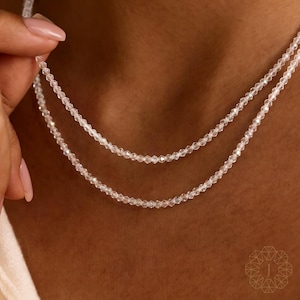 Könnte beinhalten: Nahaufnahme einer doppelreihigen Halskette aus kleinen, klaren, facettierten Perlen. Die Halskette wird von einer Hand gehalten, wodurch das filigrane Design hervorgehoben wird. Die Perlen funkeln und reflektieren das Licht. Ein hochwertiger Schmuck.