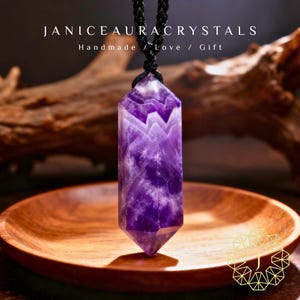 May include: A purple amethyst crystal pendant hangs from a black cord. The crystal is a deep violet color with white veining. The text "JANICEAURACRYSTALS Handmade / Love / Gift" is at the top of the image. The pendant is displayed on a wooden plate.