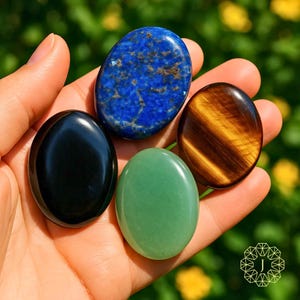 Peut inclure: Une main présente cinq pierres précieuses ovales polies. Les pierres comprennent un lapis-lazuli bleu foncé, un œil de tigre avec des rayures marron et or, une obsidienne noire et une aventurine verte. Les pierres sont lisses et réfléchissantes.