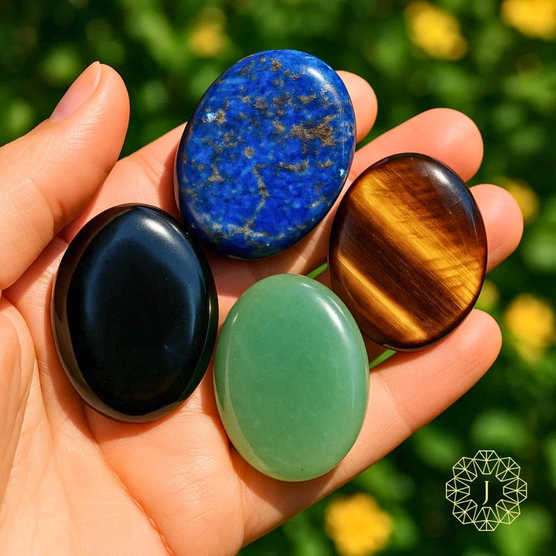 May include: A hand holds five polished, oval gemstones. The stones are a deep blue lapis lazuli, a tiger's eye with brown and gold stripes, a black obsidian, and a green aventurine. The gemstones are smooth and reflective.
