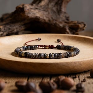 May include: A beaded bracelet with dark gray and brown stones and gold-colored accents. The bracelet is on a wooden plate with a red cord closure. The beads are approximately 0.25 inches in diameter.