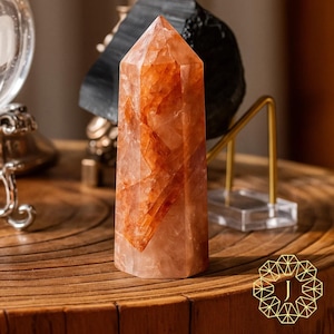 May include: A tall, orange-red crystal point with a faceted top. The crystal has internal patterns and variations in color. It is displayed on a wooden surface, with other decorative items in the background. The crystal is a decorative item.