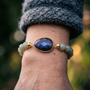 May include: A gold-toned bracelet featuring a large, faceted, teardrop-shaped, dark blue gemstone. The gemstone is the focal point, set in a gold bezel. The bracelet also includes smaller, round, pale green beads.