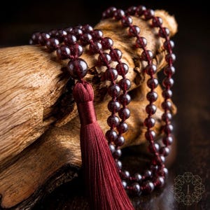 May include: A long necklace featuring deep red, translucent spherical beads strung together in a continuous loop. The necklace includes a prominent, larger central bead attached to a long, dark red silk tassel that hangs vertically from the strand.