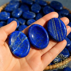 May include: Three polished, oval-shaped lapis lazuli stones in a hand, showcasing a deep blue hue with streaks of gold and white. The stones are smooth and reflective, with a basket of similar stones in the background.