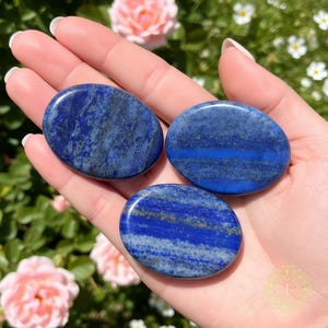 May include: Three oval-shaped lapis lazuli stones. The stones are a deep blue color with streaks of lighter blue and gold flecks. The stones are smooth and polished, and are held in a hand.