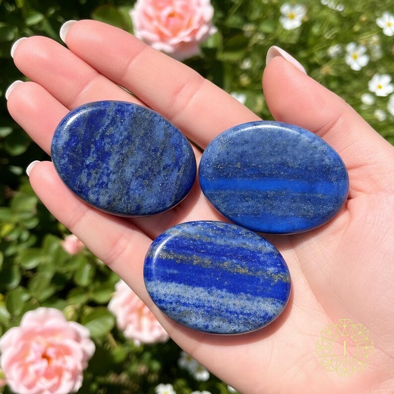 May include: Three oval-shaped lapis lazuli stones. The stones are a deep blue color with streaks of lighter blue and gold flecks. The stones are smooth and polished, and are held in a hand.