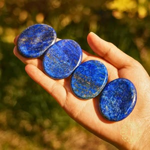 May include: Four oval-shaped lapis lazuli stones with a deep blue color and gold flecks are held in a hand. The stones are smooth and polished, with a natural, organic shape. The background is blurred, suggesting an outdoor setting.