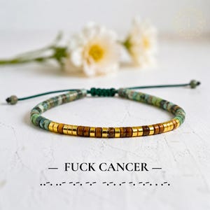 May include: A beaded bracelet featuring green mottled stone beads and a central section of alternating gold-tone and brown tiger eye beads. The bracelet has a dark green cord with an adjustable knot closure and displays the text FUCK CANCER.