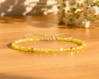 May include: A yellow beaded bracelet with gold accents. The bracelet is made of small, round, yellow beads strung together on a beige cord. The bracelet is on a wooden surface. The text "JANICEAURACRYSTALS Handcrafted Jewelry" is visible.