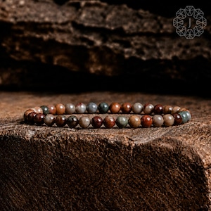 May include: A beaded bracelet featuring smooth, round stones in earthy tones of deep red, muted brown, slate gray, and forest green. The beads are strung in a continuous circular pattern, showcasing natural color variations and a polished finish.