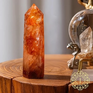 May include: A tall, orange crystal point with a faceted surface. The crystal is set on a wooden table. The crystal is a vibrant orange color with internal patterns. A smaller, clear crystal is visible in the background.