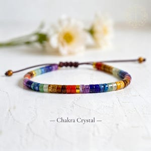 May include: A bracelet featuring small, multicolored disc-shaped beads in shades of red, orange, yellow, green, blue, and purple, arranged in a repeating pattern. The bracelet includes a dark brown cord with adjustable knots and the text Chakra Crystal.
